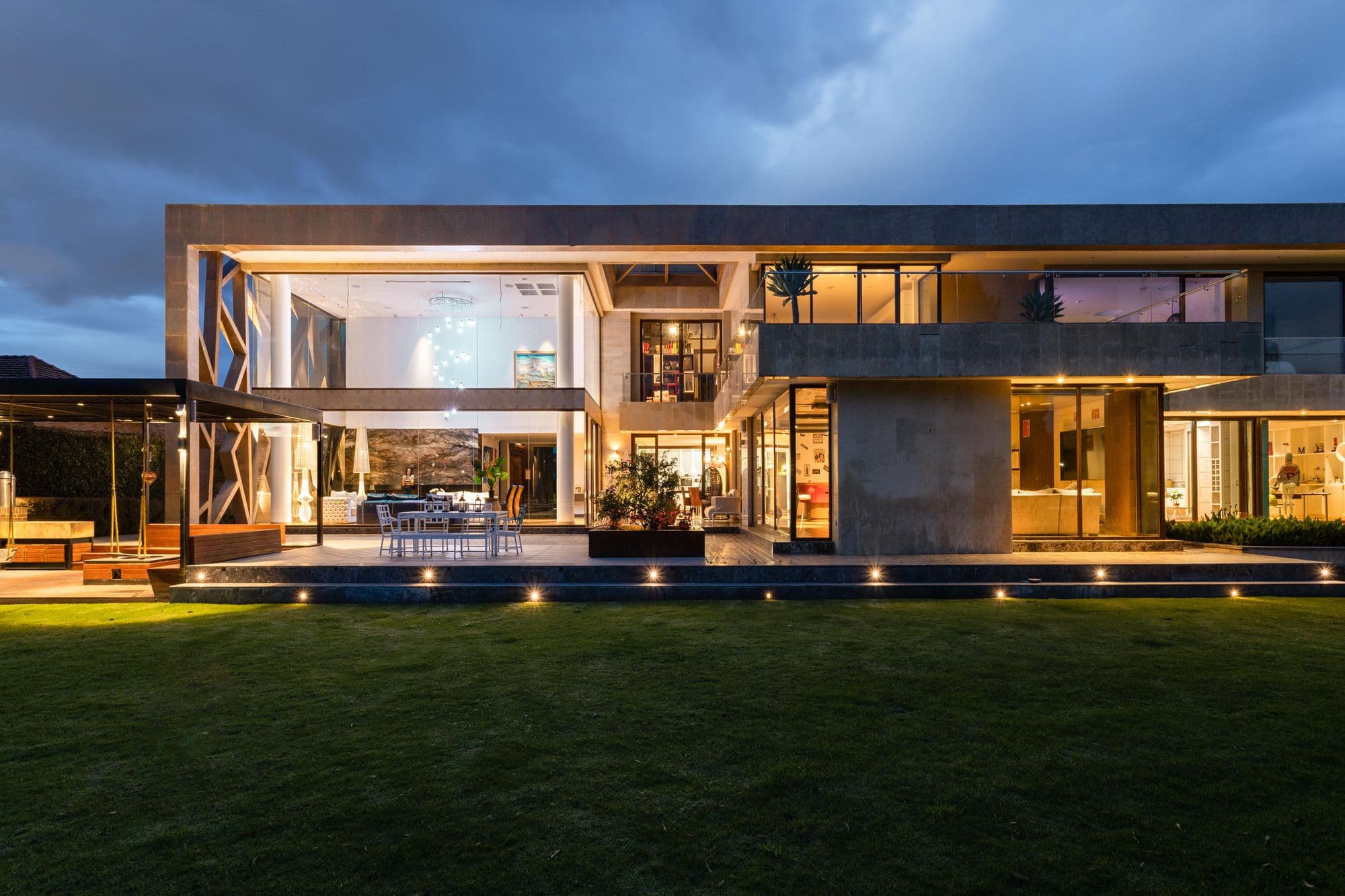 A modern luxury home with two-story floor-to-ceiling windows glowing warm at dusk over a manicured lawn